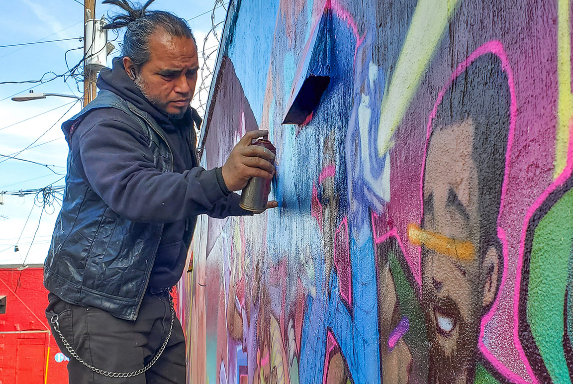 A man with salt-and-pepper hair uses a spray can to paint a colorful mural on the side of a building.