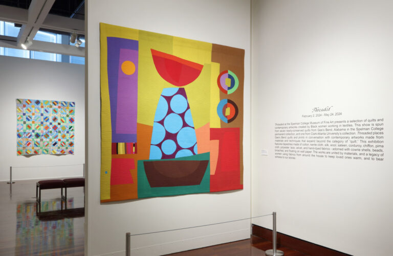 View of installation of "threaded" featuring a brightly colored modern quilt next to wall text describing the exhibition.