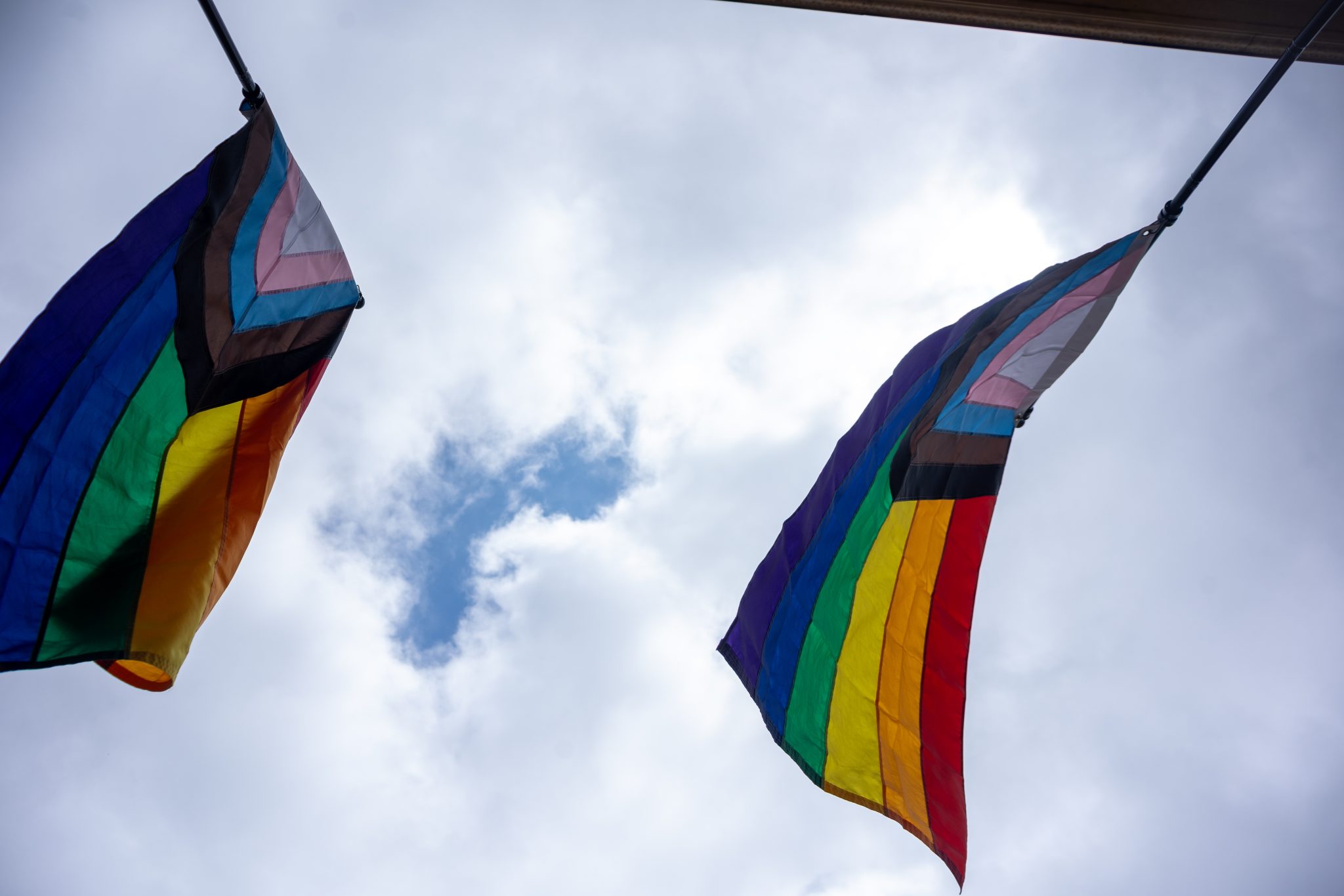 Looking up at two progress pride flags flying in the wind with just a hint of blue sky peaking out from the clouds