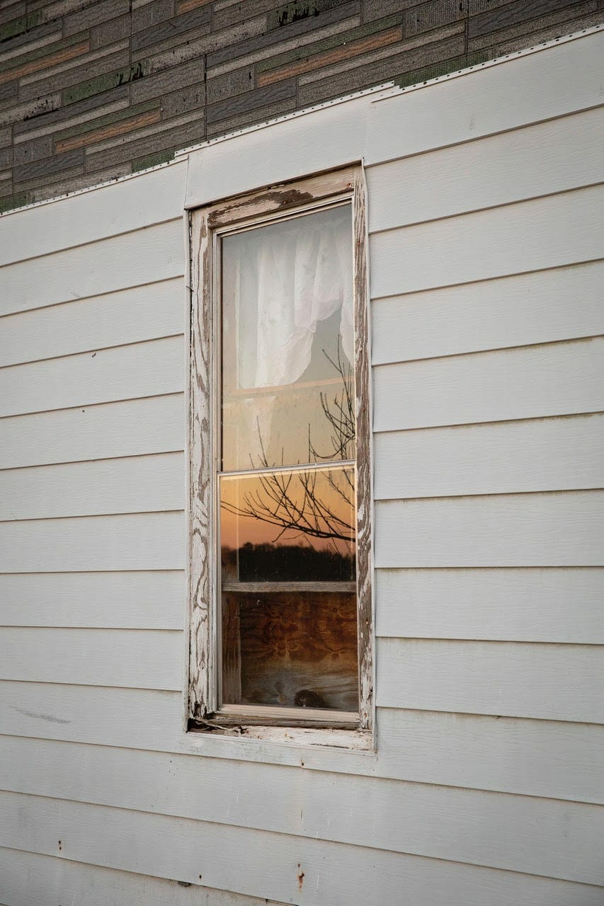 A photo by Amanda Greene of a sunset reflected in the window of an old wood-frame house.